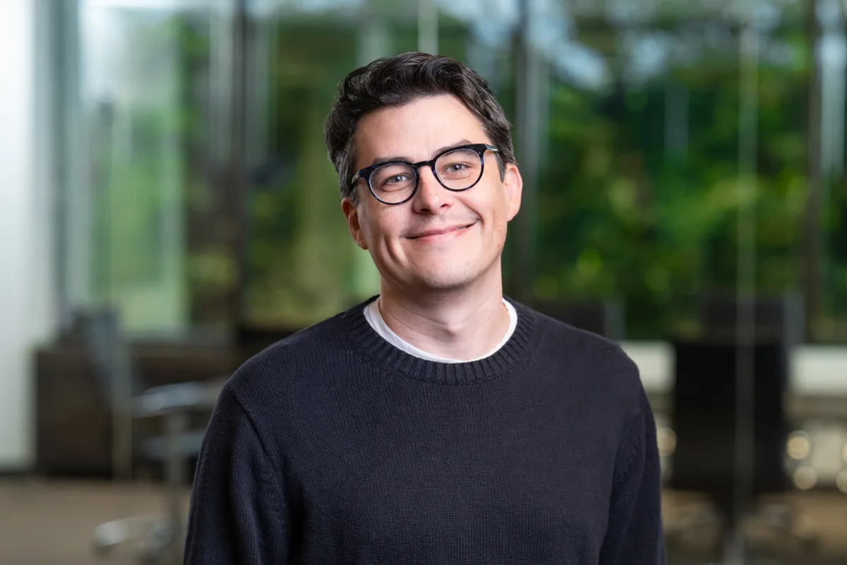 Jordan Harrison, smiling, in a dark sweater and round glasses, in front of a glass-walled office with trees behind it
