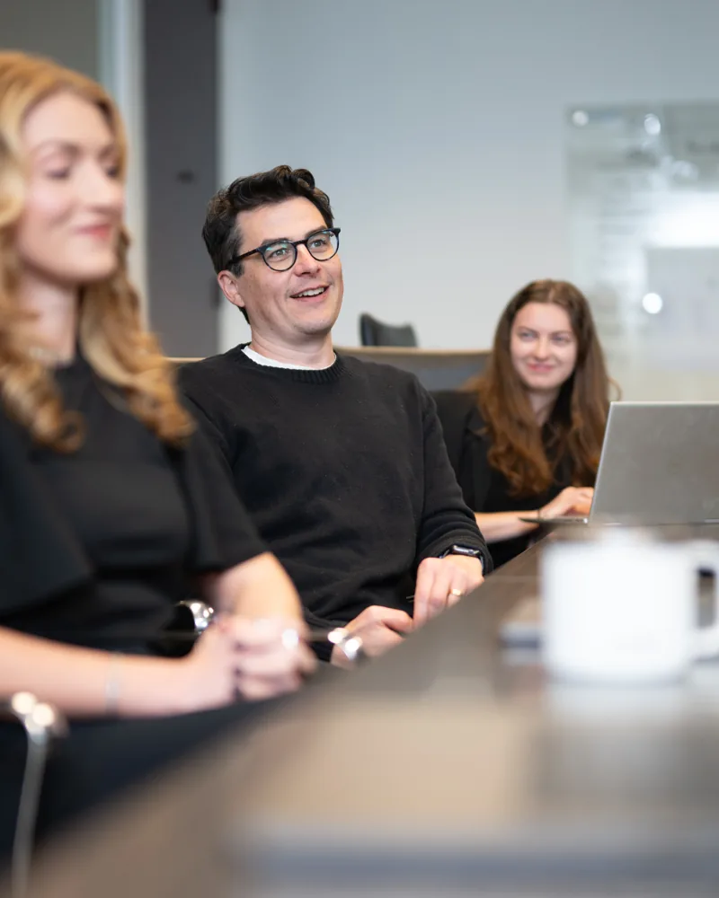 Jordan Harrison in a dark sweater and round glasses, smiling mid-conversation at a conference table, with two colleagues at laptops either side